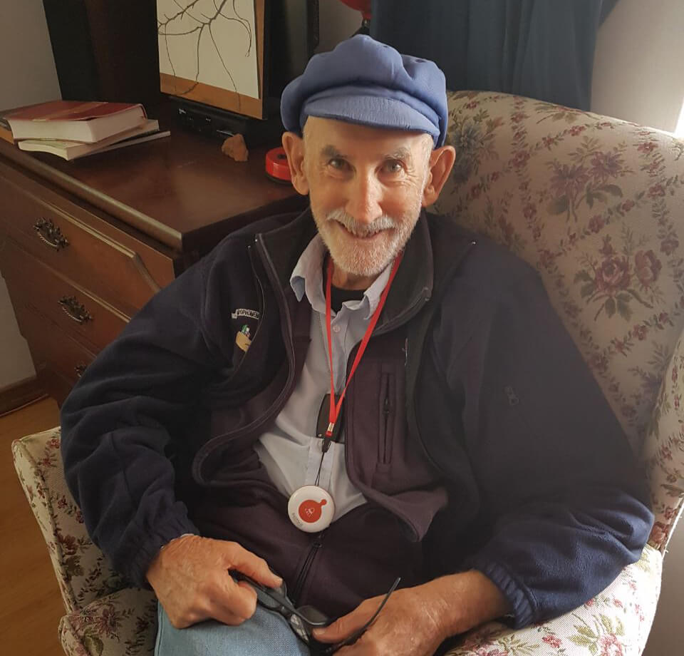 An elderly man seated at home wearing a BellMe emergency pendant on a lanyard